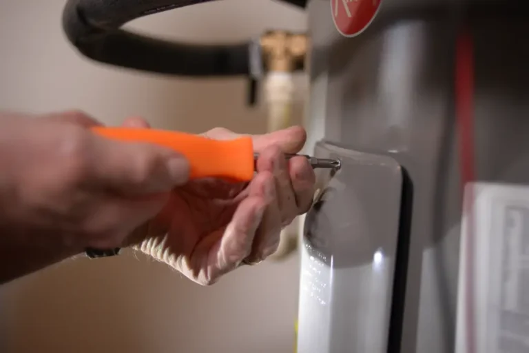 A person uses an orange-handled screwdriver to tighten or loosen a screw on a gray metal appliance.