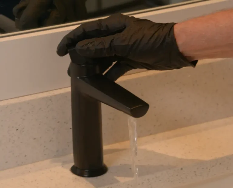 A person wearing a black glove turns on a black faucet, with water flowing into a light-colored sink.